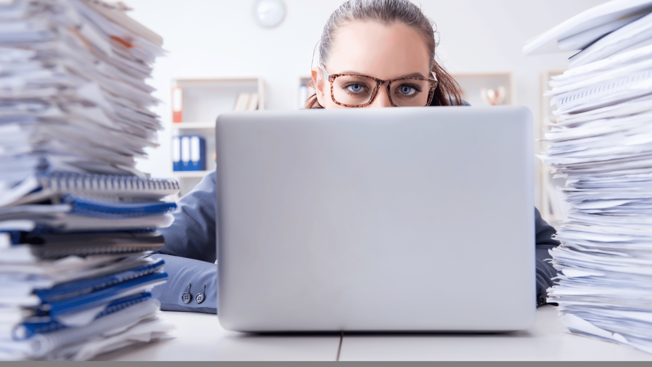 person looking up from behind a computer surrounded by paperwork